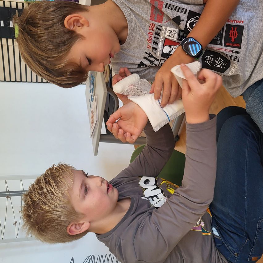 Zwei Jungen sind drinnen, einer wickelt einem anderen einen Verband um das Handgelenk. Der Junge mit dem Verband trägt ein graues Shirt und der andere trägt ein schwarzes Shirt mit verschiedenen Aufklebern.