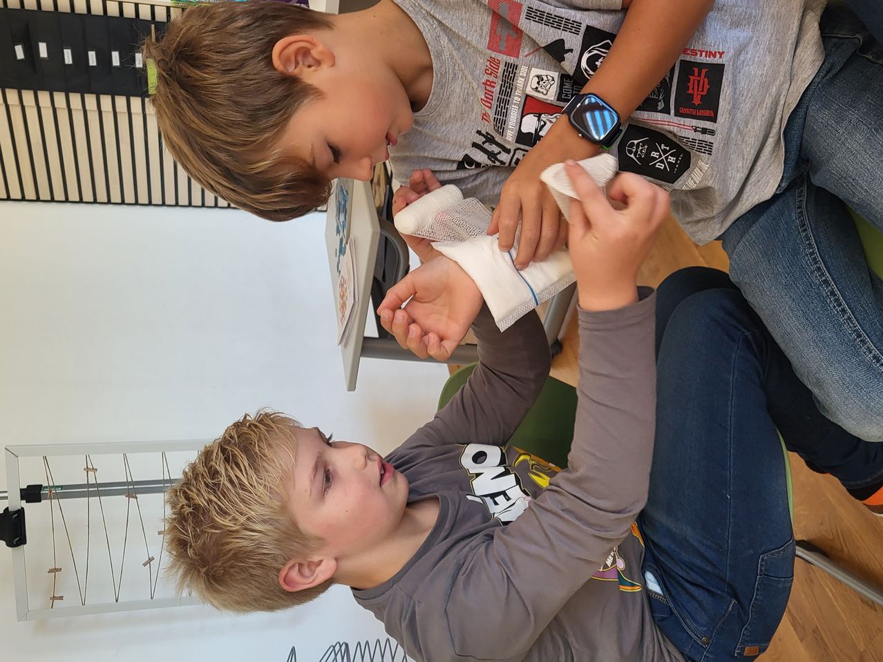 Zwei Jungen sind drinnen, einer wickelt einem anderen einen Verband um das Handgelenk. Der Junge mit dem Verband trägt ein graues Shirt und der andere trägt ein schwarzes Shirt mit verschiedenen Aufklebern.