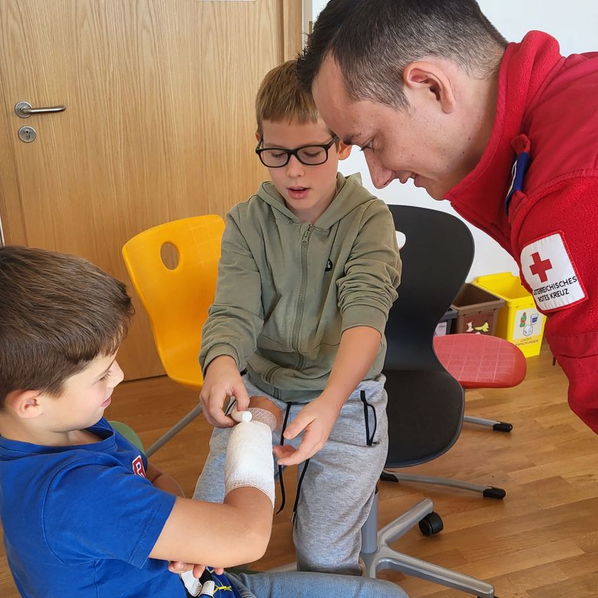 Ein Junge in einem blauen T-Shirt sitzt auf dem Boden mit einem Verband am Arm. Ein Junge mit Brille und einer grünen Kapuzenjacke schaut auf den Verband. Ein Mann in einem roten Jackenmantel mit einem weißen Kreuz hilft.