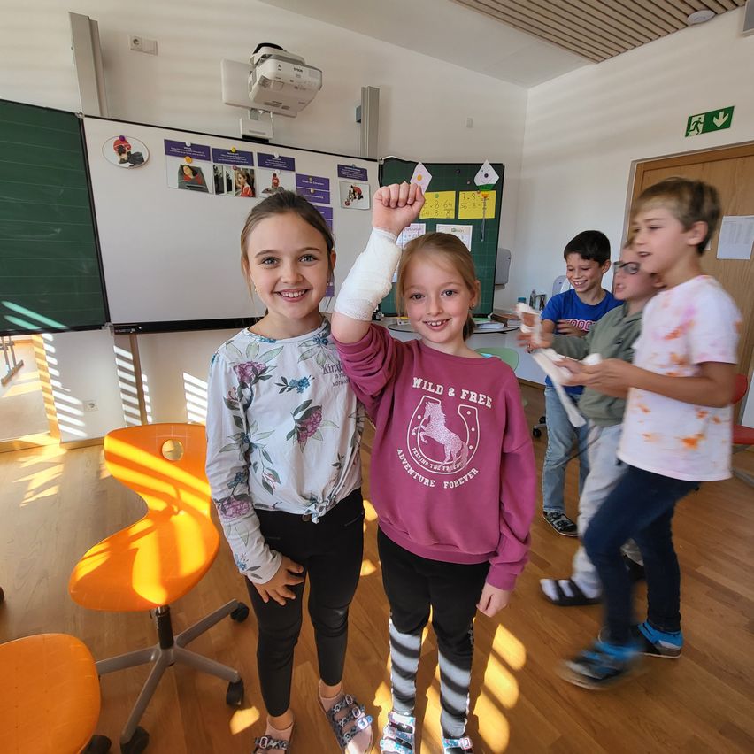 Mehrere Kinder befinden sich in einem Klassenzimmer mit einer Tafel und Stühlen. Zwei Mädchen stehen und lächeln, eines mit einem bandagierten Arm. Dahinter hält ein weiteres Kind Papier, und andere stehen und schauen sie an.