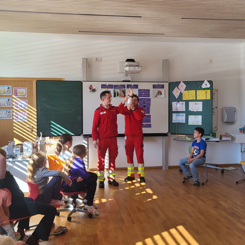 Zwei Feuerwehrleute in roten Uniformen demonstrieren etwas für eine Gruppe von Kindern, die auf Stühlen sitzen. Der Raum hat einen Holzboden und weiße Wände.