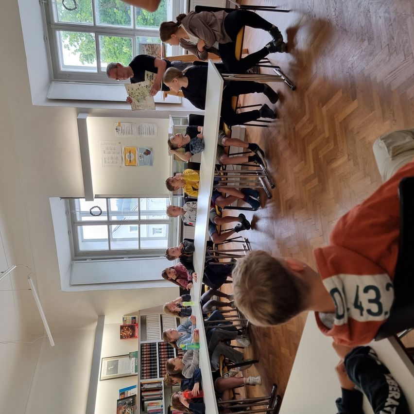 Eine Gruppe von Schülern sitzt um Tische in einem Klassenzimmer. Einige schauen in Bücher. Ein Mann steht in der Nähe eines Fensters mit Bäumen draußen.