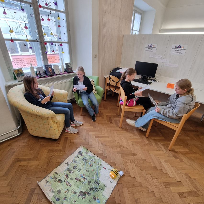 Vier Mädchen sitzen in einem Raum. Drei von ihnen lesen, während das vierte einen Laptop betrachtet. Ein Kartenausschnitt liegt auf dem Boden.