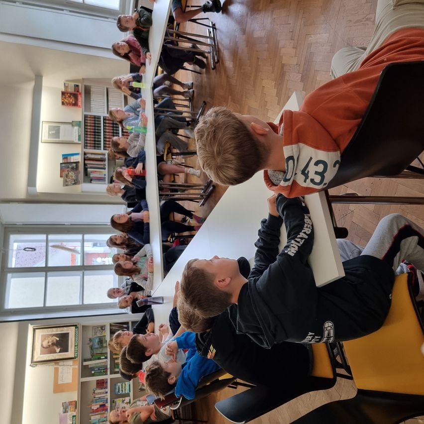 Ein Klassenzimmer mit Holzboden, in dem mehrere Schüler um Tische herum sitzen. Einige Schüler schauen zur Vorderseite. Rahmen und Bücher schmücken die Wände.