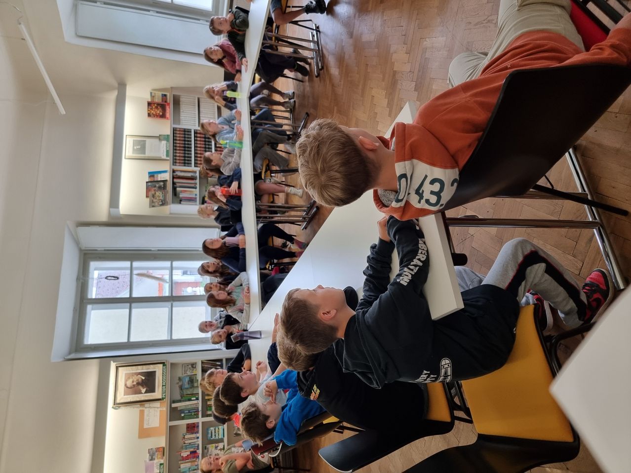 Ein Klassenzimmer mit Holzboden, in dem mehrere Schüler um Tische herum sitzen. Einige Schüler schauen zur Vorderseite. Rahmen und Bücher schmücken die Wände.