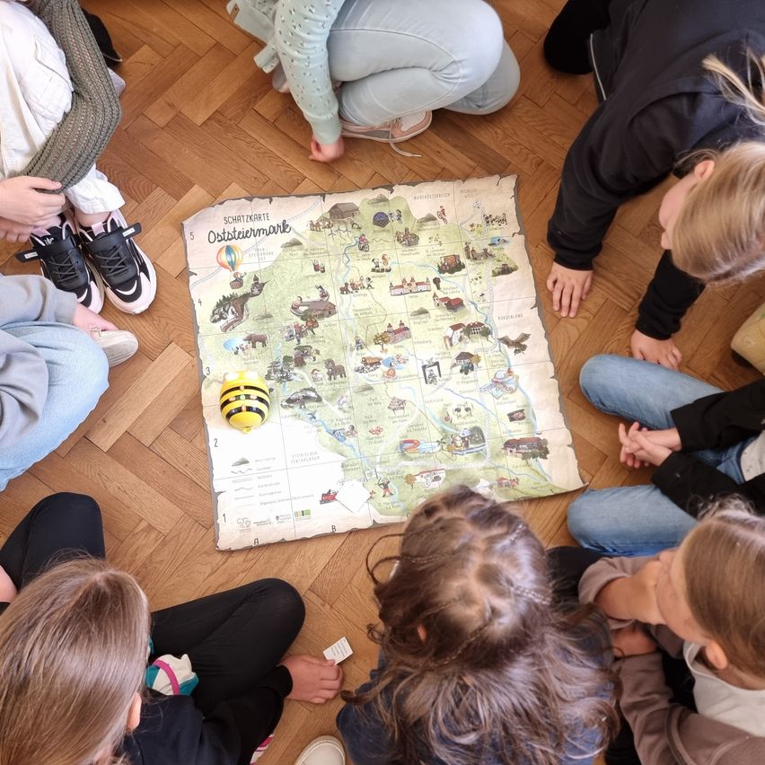 Eine Gruppe von Kindern sitzt um eine Karte auf einem Holzboden herum. Eine Spielzeugbiene ist auf der Karte. Die Kinder betrachten die Karte.
