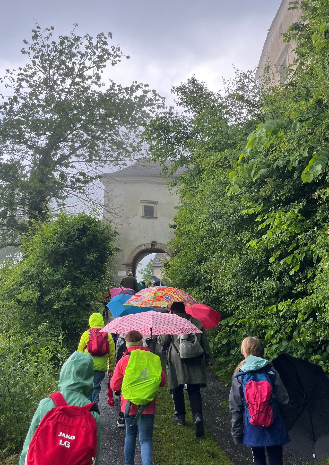 Eine Gruppe von Menschen mit Rucksäcken geht bei Regen auf einem Pfad und hält Regenschirme, mit einem Gebäude und einem Bogen im Hintergrund.