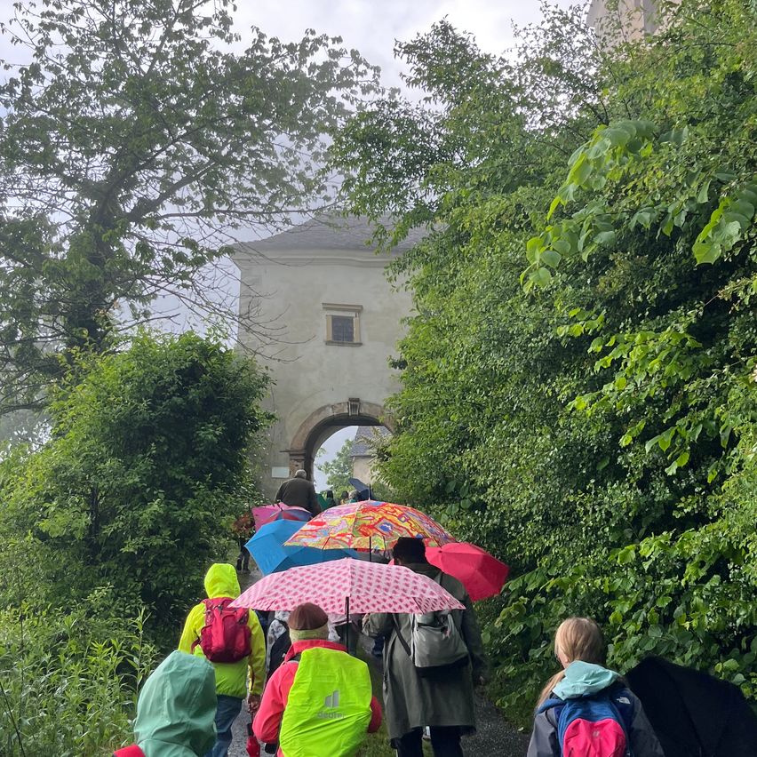Eine Gruppe von Menschen mit Rucksäcken geht bei Regen auf einem Pfad und hält Regenschirme, mit einem Gebäude und einem Bogen im Hintergrund.