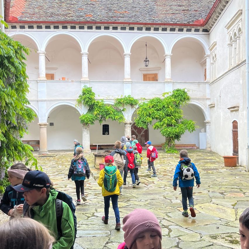 Eine Gruppe von Kindern mit Rucksäcken geht durch einen gewölbten Innenhof mit Steinboden. Der Hof ist von weißen Wänden mit gewölbten Öffnungen umgeben. Grüne Pflanzen hängen vom Bogen.