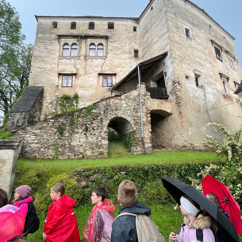 Eine Gruppe von Kindern in Regenmänteln steht vor einer alten, verwitterten Burg mit einer Steinmauer. Die Burg hat mehrere Fenster und einen gewölbten Eingang. Sie halten Regenschirme, ein Kind trägt eine weiße Stirnbinde.