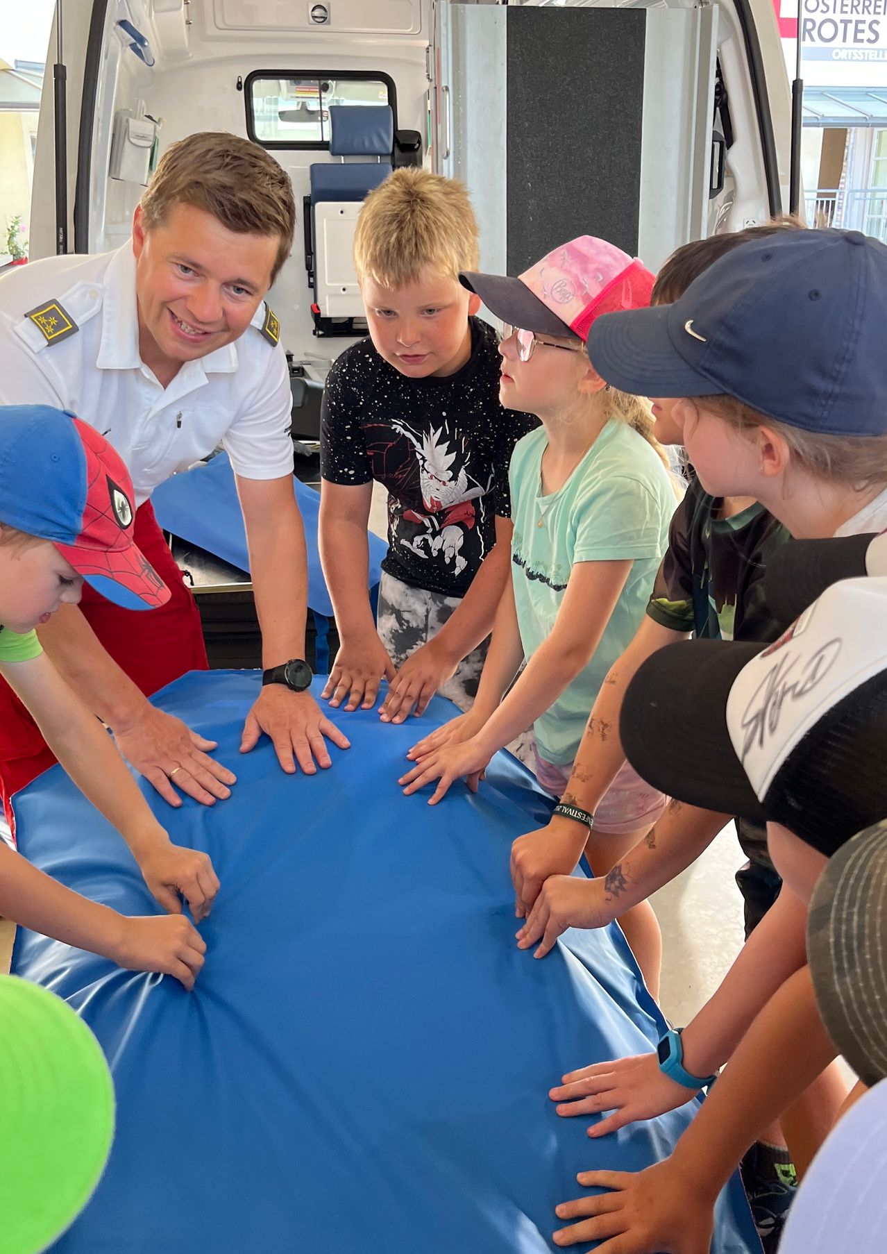 Ein lächelnder Mann in Uniform zeigt Kindern, wie man ein blaues Laken benutzt. Die Kinder tragen Mützen und schauen den Mann an.