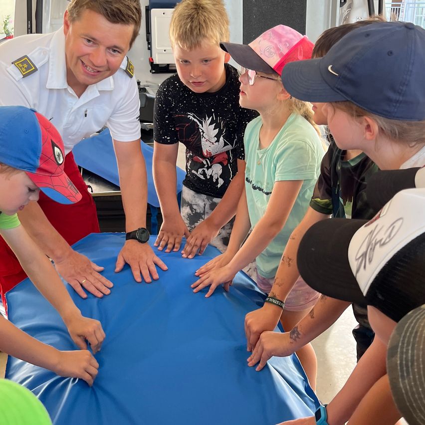 Ein lächelnder Mann in Uniform zeigt Kindern, wie man ein blaues Laken benutzt. Die Kinder tragen Mützen und schauen den Mann an.