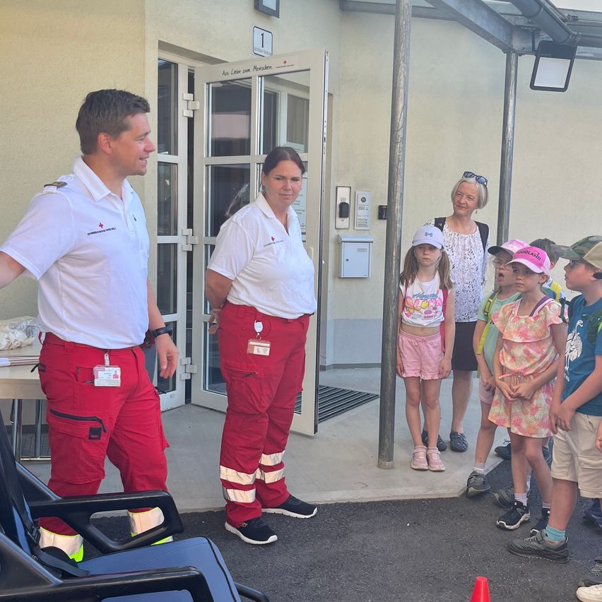 Zwei Erwachsene in weißen und roten Uniformen stehen vor einem Gebäude. Eine Gruppe von Kindern steht in der Nähe und schaut zu. Ein Erwachsener lächelt in die Kamera.