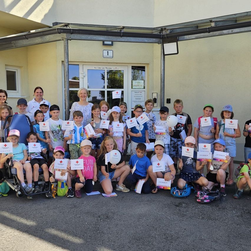 Eine Gruppe von Kindern und Erwachsenen posiert für ein Foto vor einem Gebäude mit Glastüren und Fenstern, das eine Tafel über der Tür hat. Die Kinder tragen Hüte und Turnschuhe. Einige sitzen auf dem Boden, andere stehen. Alle halten weiße Papiere mit roten Kreuzen.