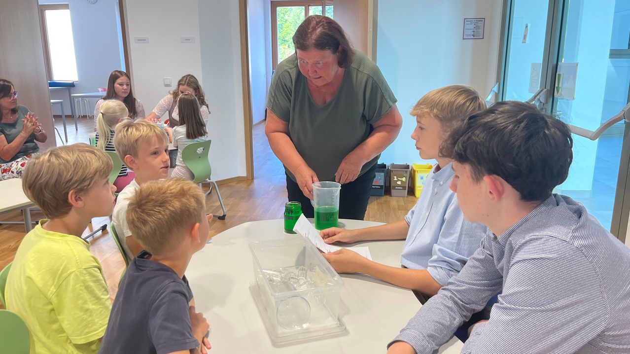 Eine ältere Frau in Grün unterrichtet Kinder bei einem wissenschaftlichen Experiment mit grüner Flüssigkeit in einem Becher auf einem Tisch. Andere Kinder beobachten.