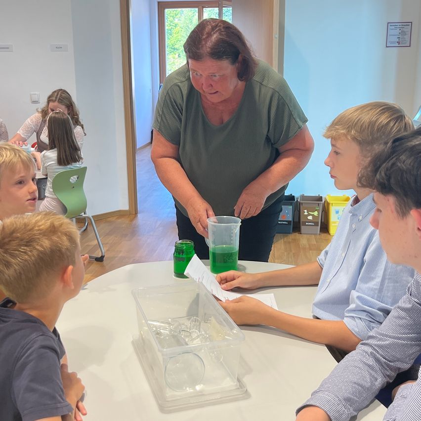 Eine ältere Frau in Grün unterrichtet Kinder bei einem wissenschaftlichen Experiment mit grüner Flüssigkeit in einem Becher auf einem Tisch. Andere Kinder beobachten.