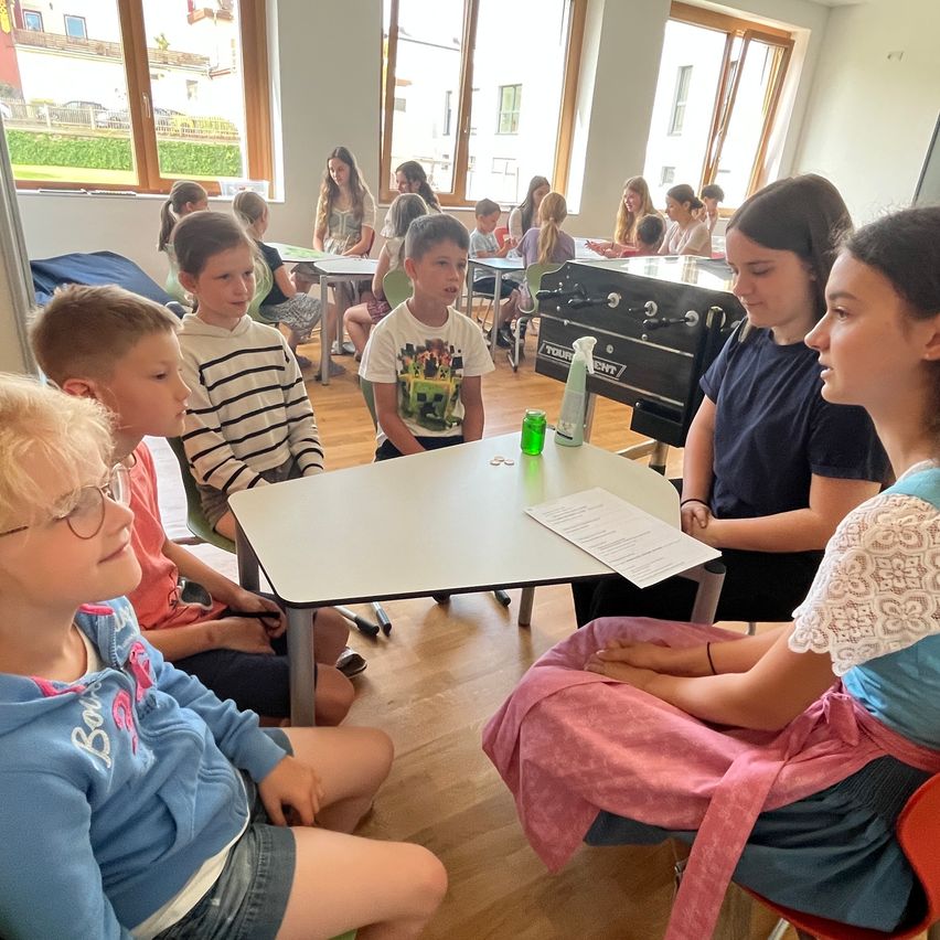 Ein Klassenzimmer mit Kindern, die um einen Tisch herum sitzen, wobei zwei Mädchen einem Jungen zuhören, der spricht. Ein Tischfußball und ein Taschentuchspender befinden sich auf dem Tisch.