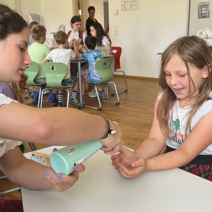 Ein junges Mädchen trägt Lotion auf die Hände eines anderen Mädchens in einem Klassenzimmer auf. Mehrere Kinder sitzen im Hintergrund an Tischen.