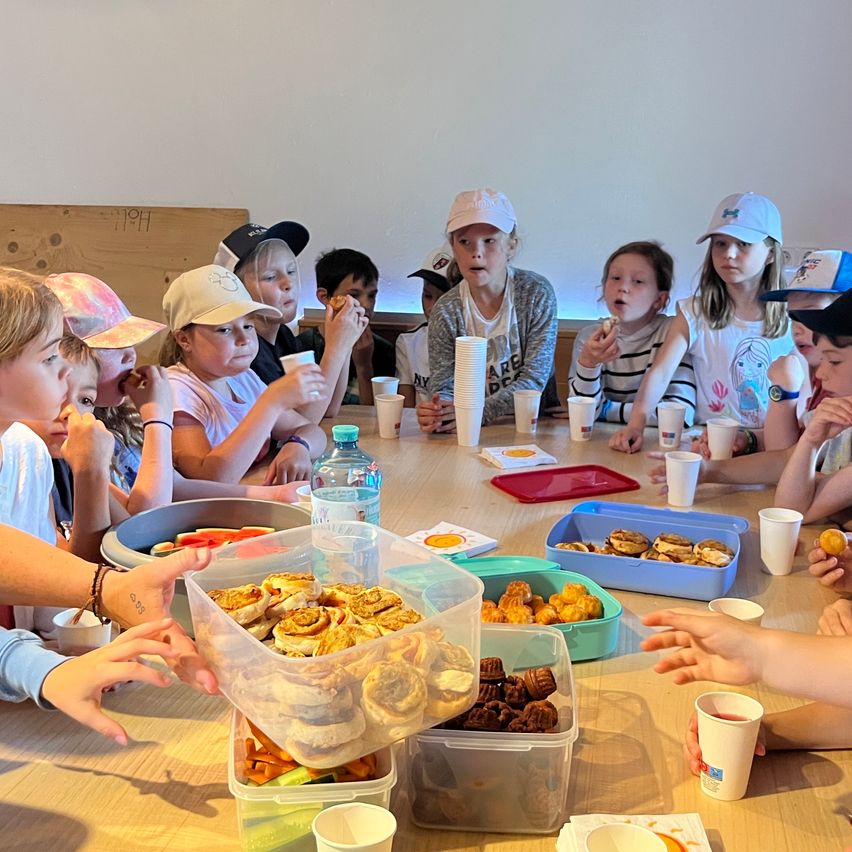 Eine Gruppe von Kindern sitzt um einen Tisch und genießt verschiedene Speisen. Eine Person verteilt Brötchen in einem durchsichtigen Behälter, während andere Kekse und Obst essen. Auf dem Tisch stehen Becher, Teller und Flaschen.