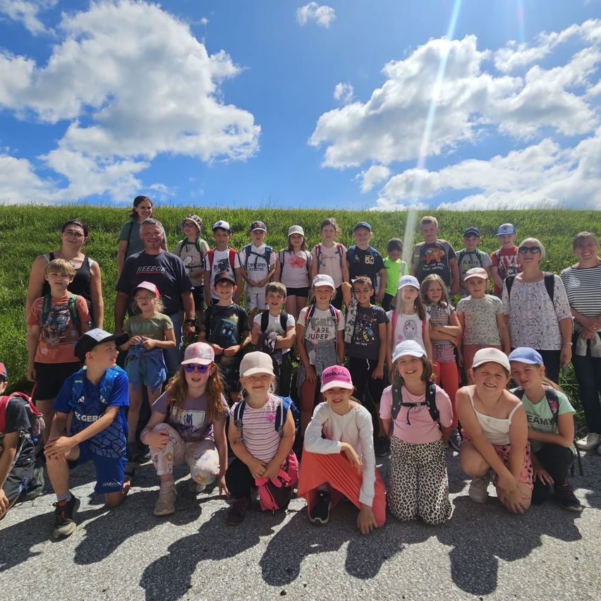 Eine Gruppe von Kindern und Erwachsenen posiert für ein Foto vor einem grün bewachsenen Hügel unter einem blauen Himmel mit Wolken.