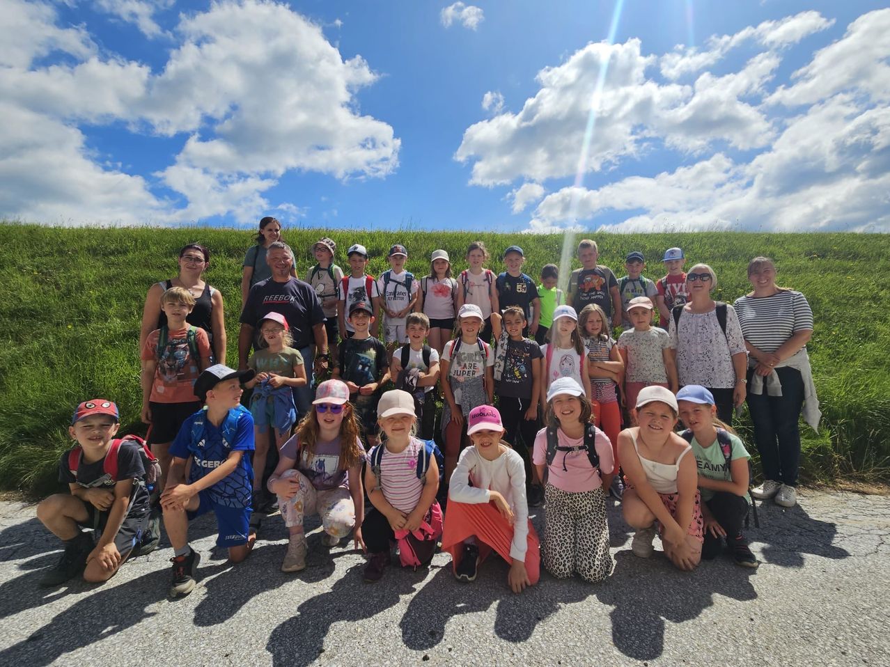 Eine Gruppe von Kindern und Erwachsenen posiert für ein Foto vor einem grün bewachsenen Hügel unter einem blauen Himmel mit Wolken.