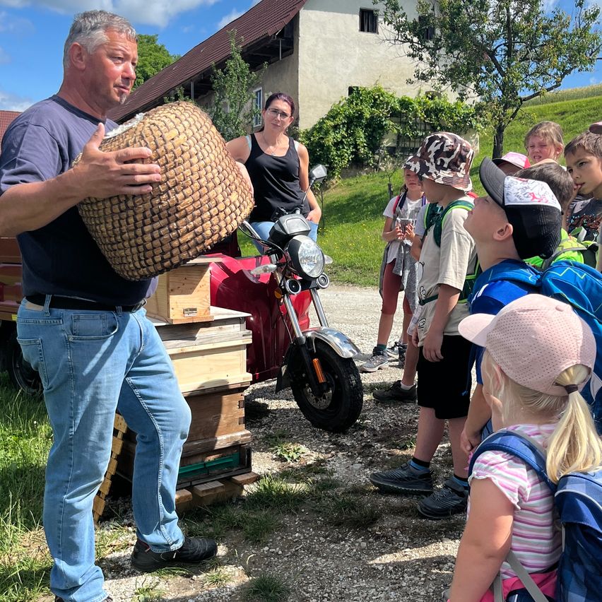 Ein Mann zeigt einer Gruppe von Kindern einen geflochtenen Korb, mit Holzbienenstöcken und einem Motorrad im Hintergrund.