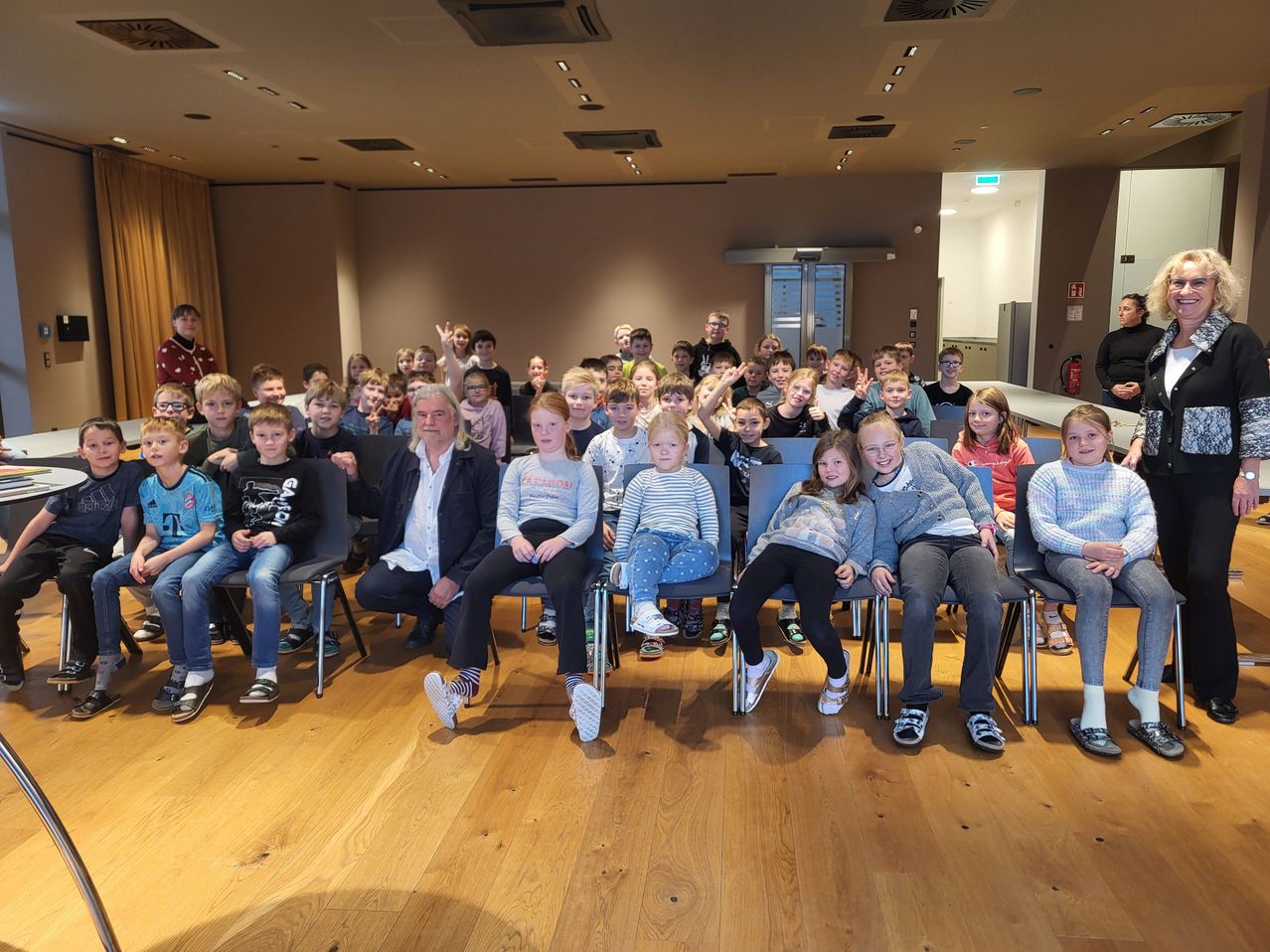 Eine Gruppe von Kindern und einem erwachsenen Mann sitzen auf Stühlen und posieren für ein Foto in einem Raum mit Holzboden und weißer Decke.