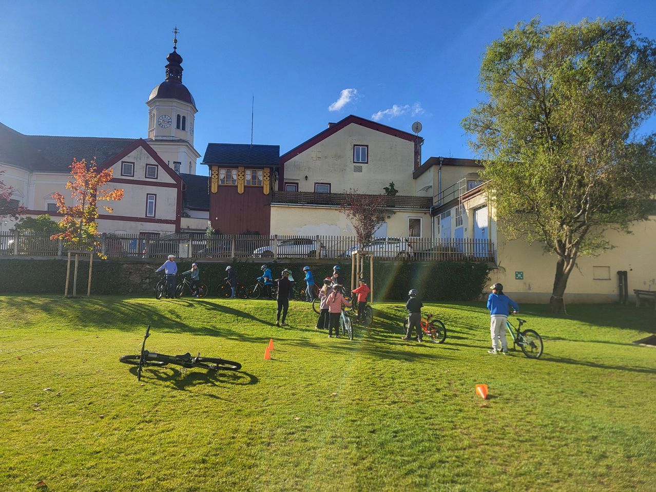 Eine Gruppe von Menschen fährt auf einem Grasfeld mit Gebäuden im Hintergrund auf Fahrrädern. Einige stehen und andere sitzen auf ihren Fahrrädern. Bäume und ein Turm sind sichtbar.