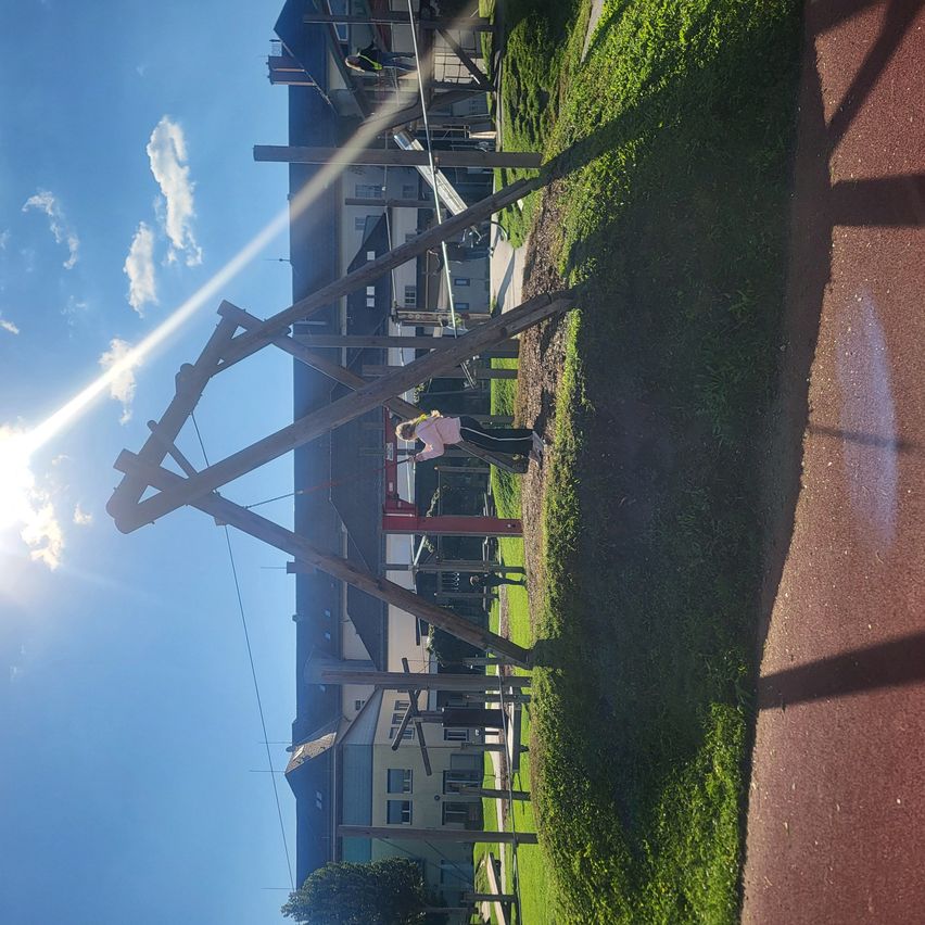 Ein Spielplatz mit einer großen Holzstruktur, einer Rutsche und einer Schaukel unter einem strahlend blauen Himmel mit ein paar Wolken. Eine Person steht im Gras.