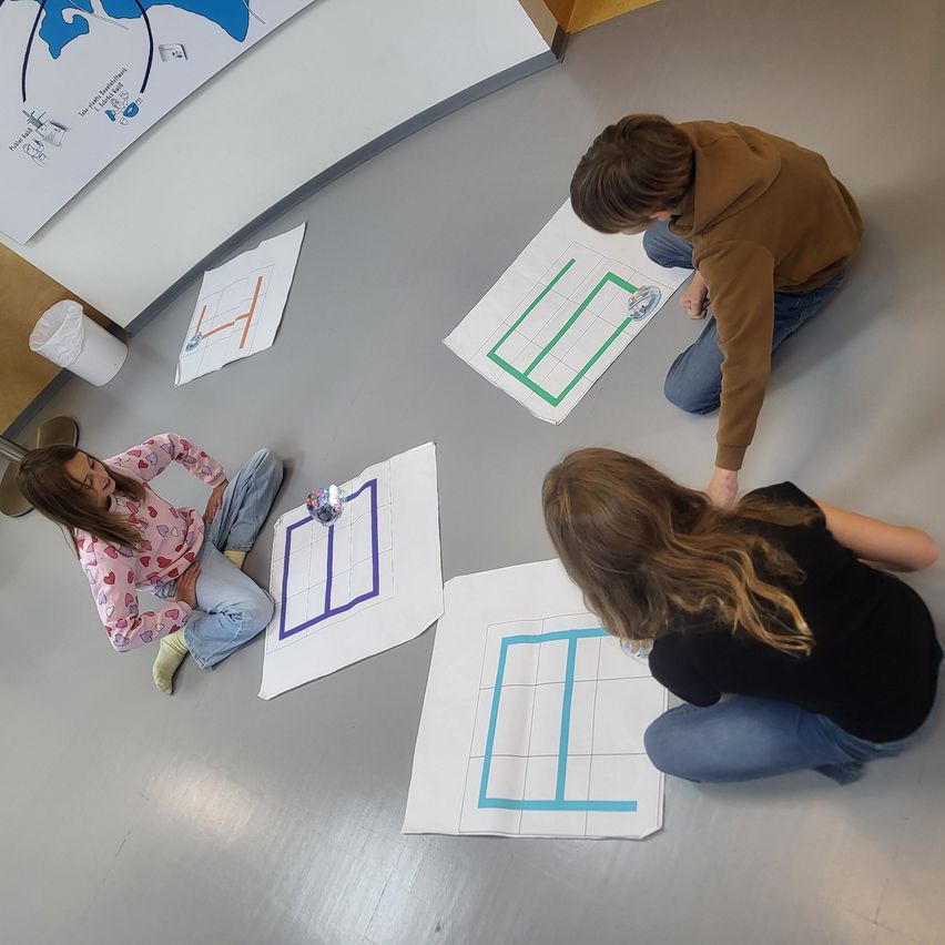 Vier Schüler sitzen auf dem Boden und arbeiten an einem Projekt. Jeder hat ein großes Stück Papier mit einem Rastermuster. Ein Junge kniet und konzentriert sich auf sein Papier.