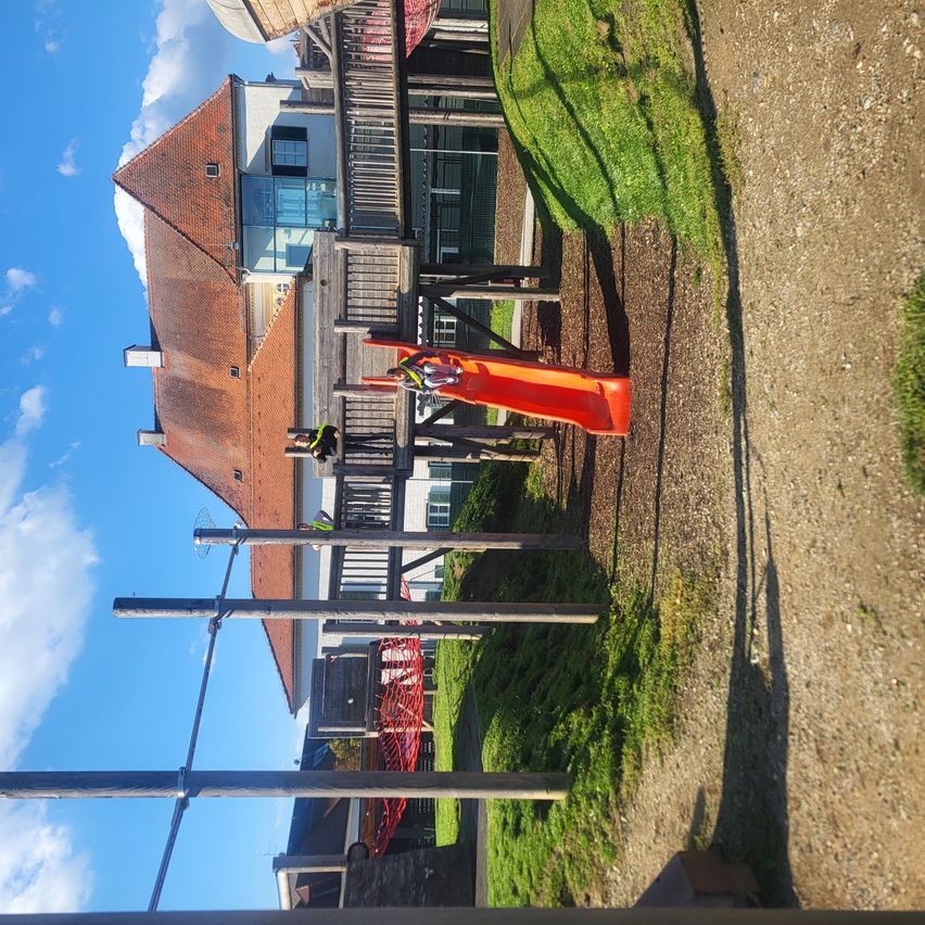 Ein Spielplatz mit einem roten Rutschen und Arbeitern in Sicherheitsausrüstung. Im Hintergrund befindet sich ein Ziegelgebäude mit Glasfenstern unter einem klaren blauen Himmel.