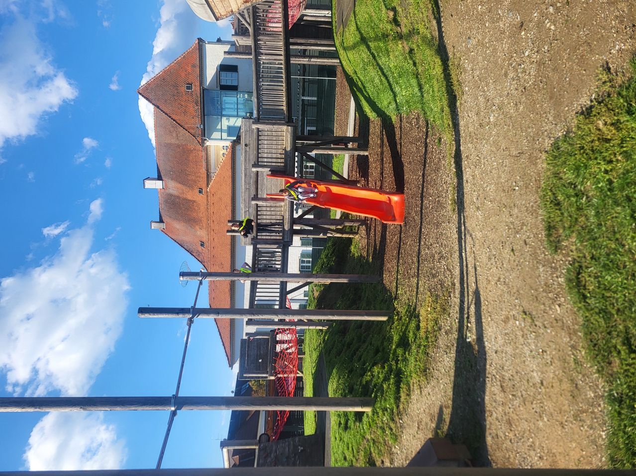 Ein Spielplatz mit einem roten Rutschen und Arbeitern in Sicherheitsausrüstung. Im Hintergrund befindet sich ein Ziegelgebäude mit Glasfenstern unter einem klaren blauen Himmel.