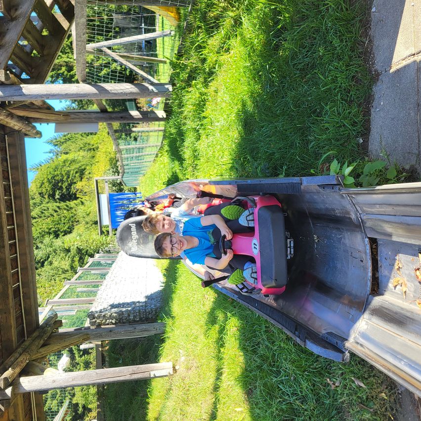 Zwei Kinder fahren mit einem Bobschlitten eine Strecke hinunter. Sie tragen Helme und Schutzbrillen. Die Strecke ist von einem Holzzaun umgeben.