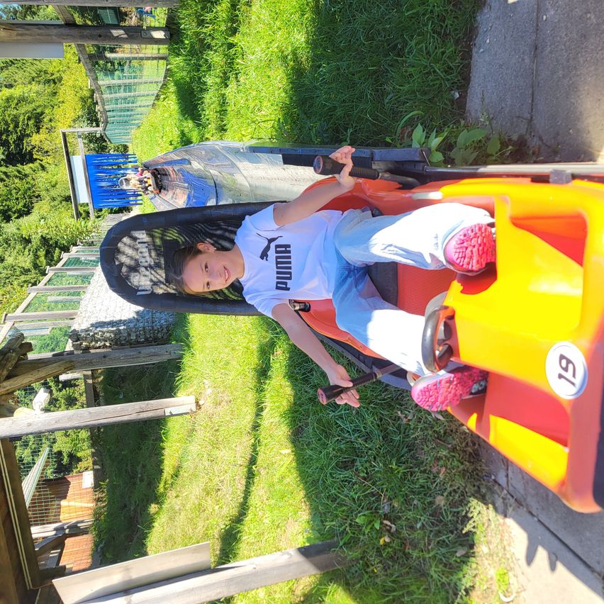 Ein junges Mädchen in einem Puma-Shirt fährt einen orange-gelben Go-Kart auf einem grasbewachsenen Pfad. Dahinter ist ein weiterer Go-Kart zu sehen. Bäume und ein Holzzaun umgeben das Gebiet.