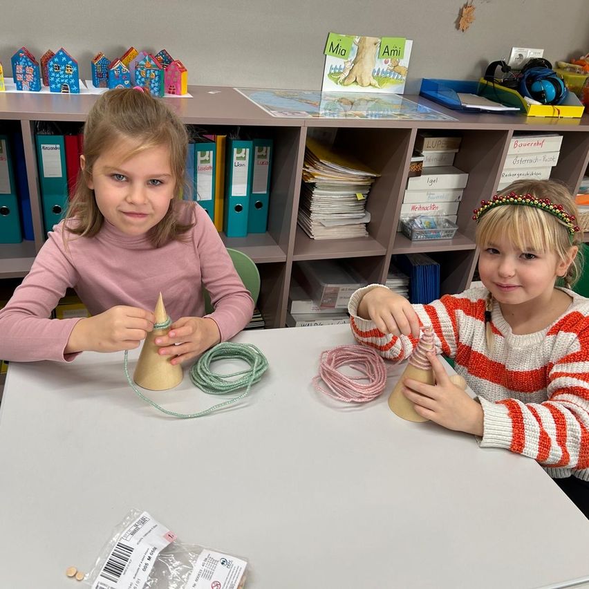Zwei junge Mädchen arbeiten an einem Bastelprojekt am Tisch. Eine hält einen Kegel, die andere einen Draht. Hinter ihnen steht ein Bücherregal mit Büchern und Ordnern.