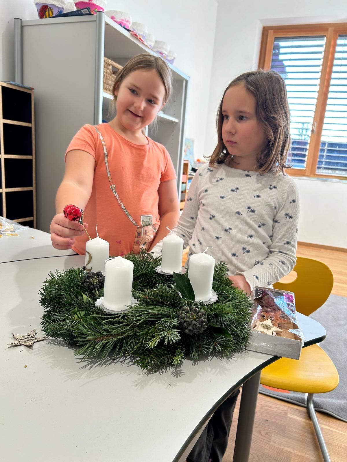 Zwei Mädchen stehen an einem Tisch mit einem Kranz aus Tannennadeln und vier brennenden Kerzen. Ein Mädchen hält eine rote Kugel und ein Karton mit Kugeln steht neben dem Kranz. Dahinter befinden sich ein Bücherregal und ein Fenster.