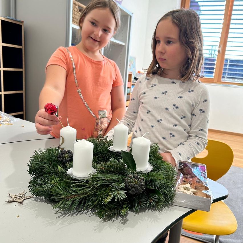 Zwei Mädchen stehen an einem Tisch mit einem Kranz aus Tannennadeln und vier brennenden Kerzen. Ein Mädchen hält eine rote Kugel und ein Karton mit Kugeln steht neben dem Kranz. Dahinter befinden sich ein Bücherregal und ein Fenster.