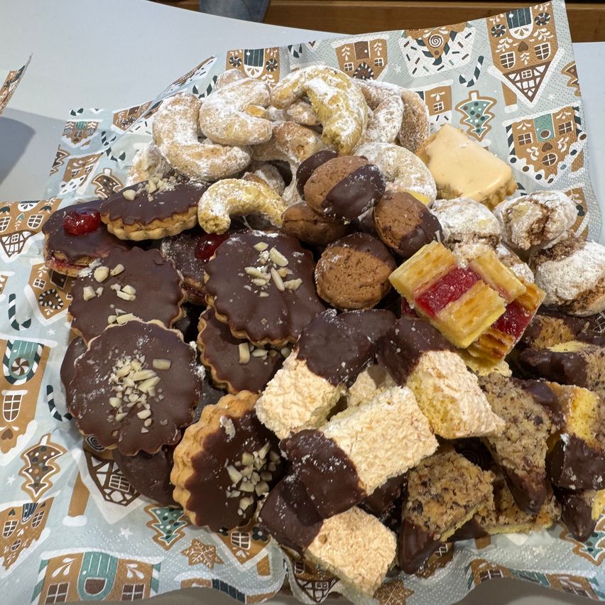 Eine festliche Auswahl an verschiedenen Gebäckstücken und Keksen, darunter schokoladenüberzogene und mit Früchten gefüllte Leckereien, liegt auf einem dekorierten Papier. Die Leckereien sind in einem Kreismuster auf einer festlichen Tischdecke angeordnet.