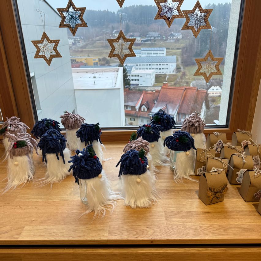 Ein Holztisch hält handgefertigte Gnome mit weißen Bärten und blauen Mützen. Ein großes Fenster dahinter zeigt eine Stadtlandschaft. Sternendekorationen hängen am Fenster.