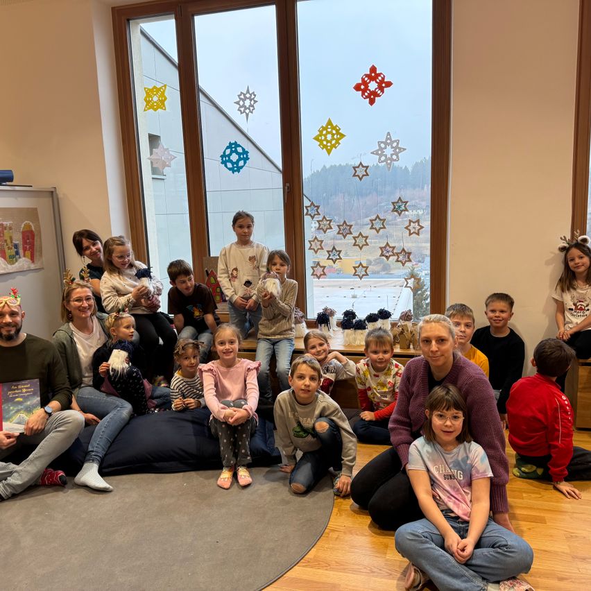 Eine Gruppe von Kindern und Erwachsenen in einem Raum mit großen Fenstern, die mit Schneeflocken verziert sind, lächeln für ein Foto. Einige Kinder sitzen auf Kissen auf dem Boden, und zwei Erwachsene stehen mit Büchern.