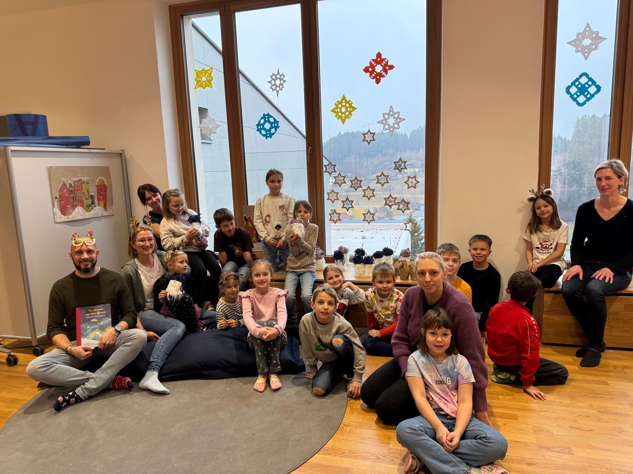 Eine Gruppe von Kindern und Erwachsenen in einem Raum mit großen Fenstern, die mit Schneeflocken verziert sind, lächeln für ein Foto. Einige Kinder sitzen auf Kissen auf dem Boden, und zwei Erwachsene stehen mit Büchern.