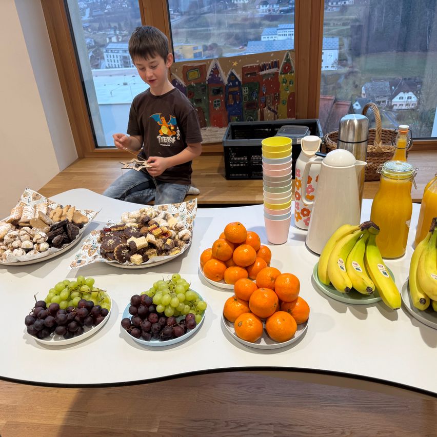 Ein junger Junge sitzt am Fenster und schaut auf einen Tisch voller Essen, einschließlich Trauben, Orangen, Bananen und Kekse.