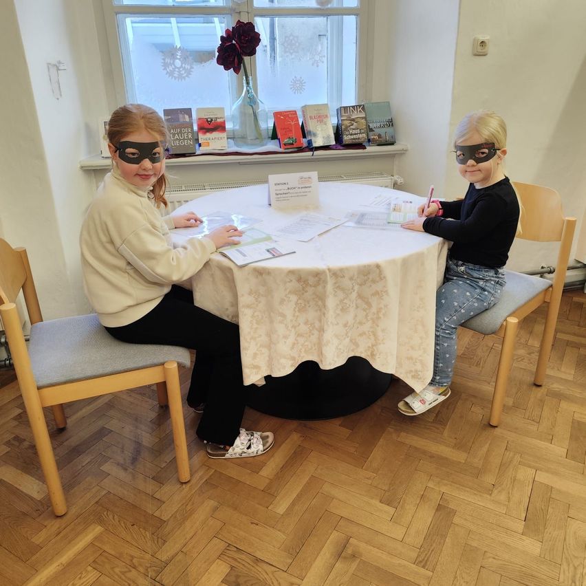 Zwei junge Mädchen mit Masken sitzen an einem runden Tisch mit Büchern und Papieren. Sie schreiben mit Stiften. Ein Fenster mit Büchern und Blumen im Hintergrund.