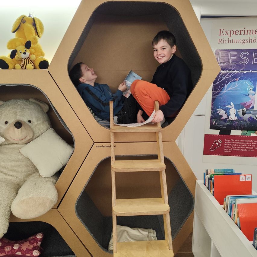 Zwei junge Jungen sitzen in einer sechseckigen Pappstruktur, lächeln und lesen ein Buch. Eine Leiter führt zum Eingang. Gefüllte Tiere und Bücher umgeben sie. Ein Plakat ist an der Wand.