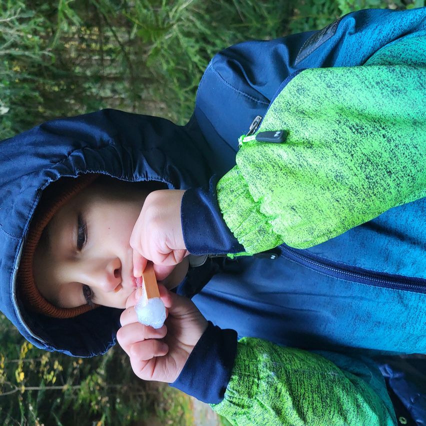 Ein Kind in einem blau- und grünfarbenen Mantel bläst auf ein Stück Eis, während es draußen ist.