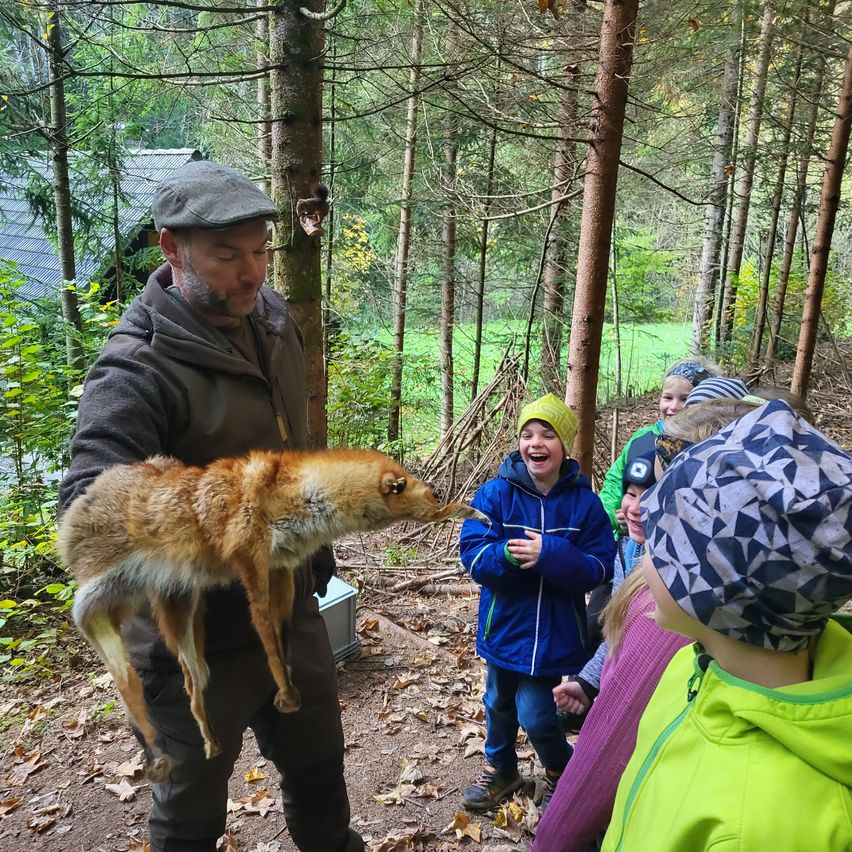 Ein Mann hält einen Kojoten in einem Wald, um ihn herum stehen Kinder, einige in Mützen, lächeln und schauen den Kojoten an.