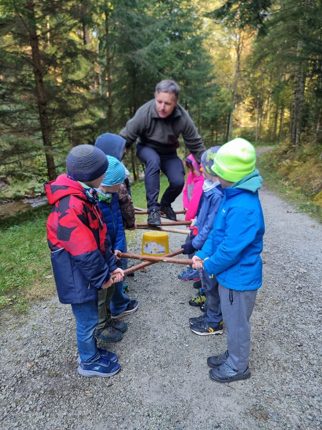 Ein Mann hilft mehreren Kindern, einen Baumstamm in einem Wald zu ziehen. Die Kinder sind in Jacken und Mützen gekleidet.
