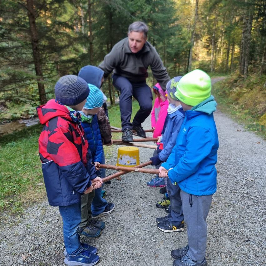Ein Mann hilft mehreren Kindern, einen Baumstamm in einem Wald zu ziehen. Die Kinder sind in Jacken und Mützen gekleidet.