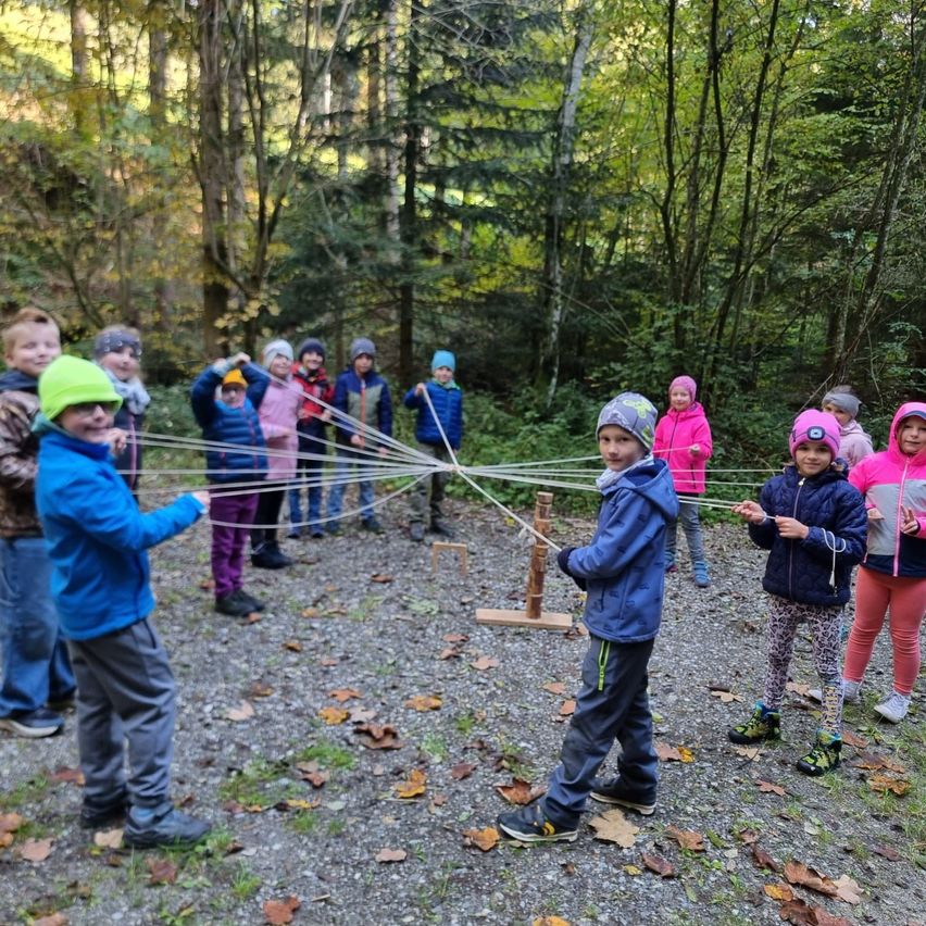 Kinder in Winterkleidung spielen mit einem Schnurspiel auf einem Schotterweg in einem Wald. Sie halten die Schnüre und sind von gefallenen Blättern und Bäumen umgeben.
