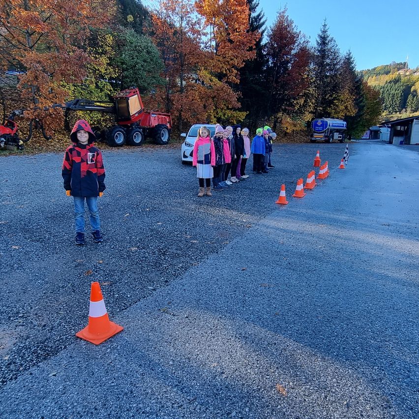 Eine Gruppe von Menschen steht auf einer Straße, die mit orangefarbenen Verkehrskegel gesäumt ist. Ein Junge steht allein, während andere hinter ihm stehen. Ein LKW und ein Auto sind auf der linken Seite geparkt. Bäume mit Herbstlaub umgeben das Gebiet.